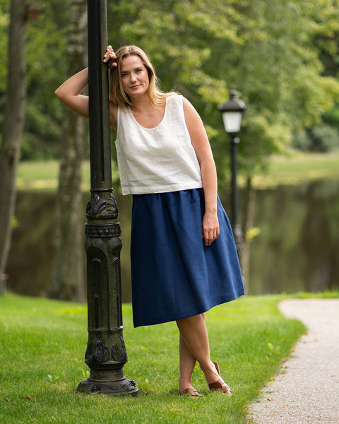 A woman rests casually against an ornate black lamppost in a lush green park with a tranquil lake in the background. She models a sleeveless pure white linen top and a storm blue midi skirt, completing the relaxed outfit with brown sandals.