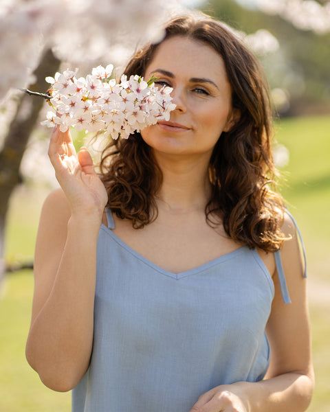 Una mujer que lleva un vestido lencero de lino azul nublado está junto a un árbol cubierto de flores blancas y huele suavemente un racimo de flores.