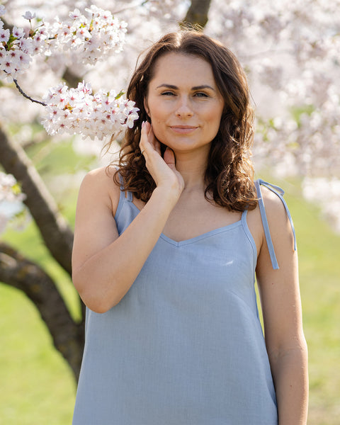 Una mujer que lleva un vestido lencero de lino azul nublado está junto a un árbol cubierto de flores blancas.