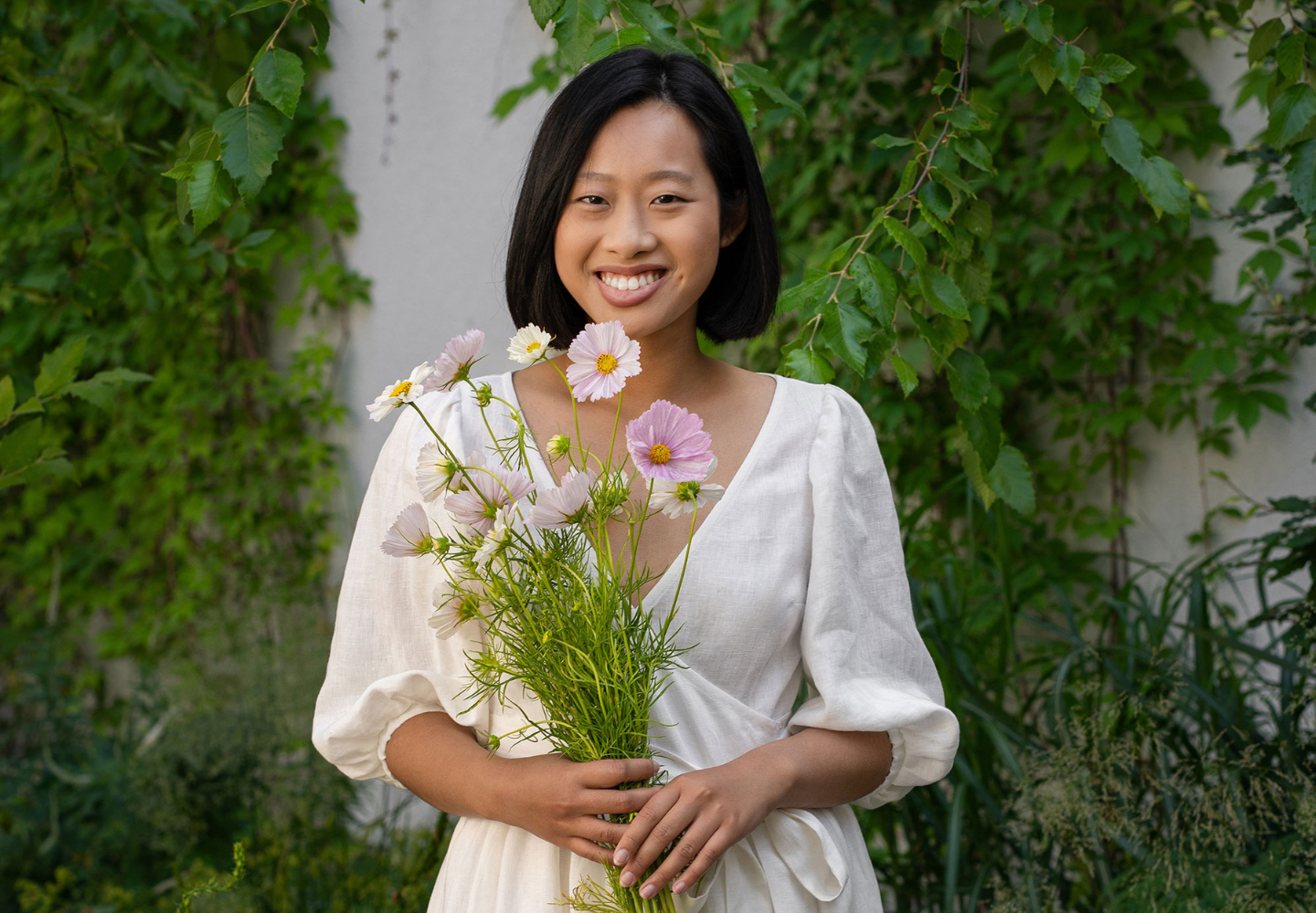Una mujer sonriente con un vestido blanco de lino de menique sostiene un ramo de flores cosmos en tonos pastel, de pie frente a un muro verde cubierto de hojas.