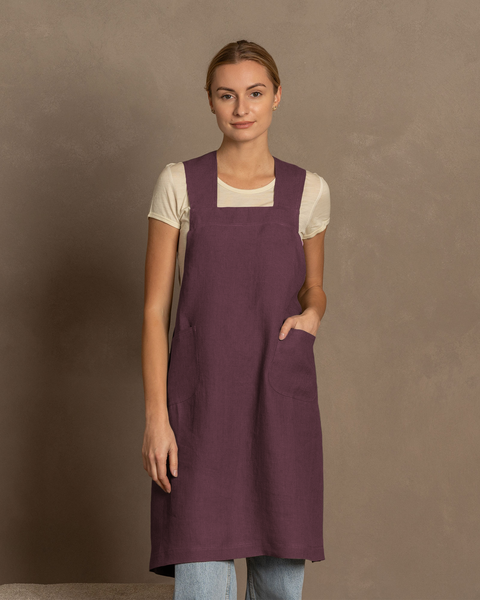 Una mujer de piel clara con cabello rubio recogido mira directamente al espectador. Lleva una camisa blanca de manga corta bajo un delantal cruzado de lino color púrpura sombra con bolsillos delanteros. Se ven unos vaqueros azul claro debajo del delantal. Está de pie frente a un fondo liso de color marrón cálido.
