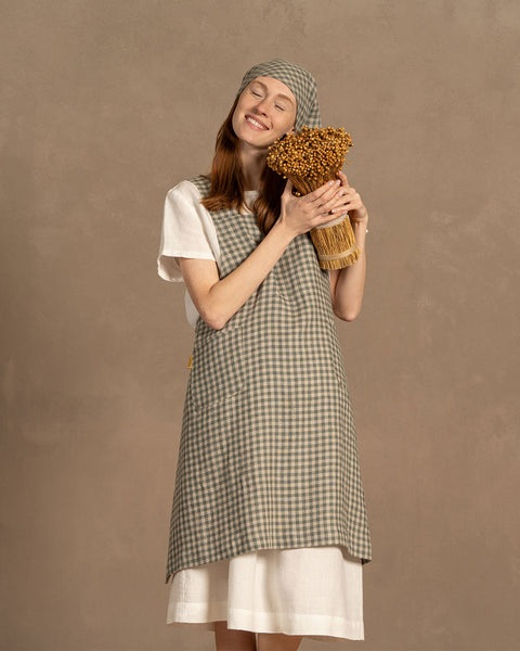 Una persona de piel clara y cabello largo castaño rojizo sonríe con los ojos cerrados, sosteniendo suavemente un ramo de flores secas doradas contra su pecho. Lleva una camisa blanca de lino de manga corta bajo un delantal verde a cuadros y un pañuelo a juego atado bajo la barbilla. El fondo es de un marrón cálido y apagado.