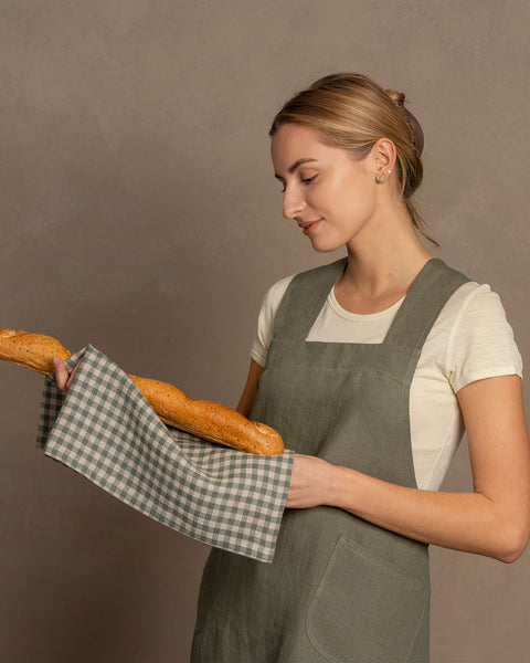 Una mujer con cabello rubio sostiene una baguette envuelta en un paño de lino de menique, vistiendo un delantal cruzado de lino verde piedra, sobre un fondo neutro.