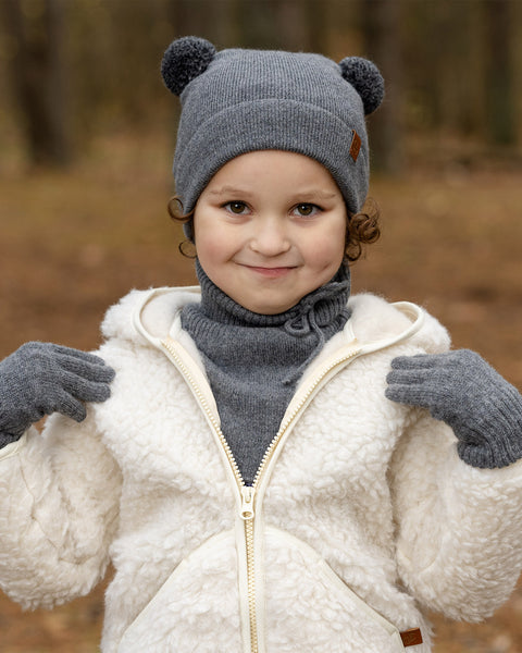 Un niño pequeño con cabello rizado castaño claro y una sonrisa sutil, mira directamente al espectador. Lleva un gorro de punto gris oscuro con dos orejas de pompón y una etiqueta marrón, mitones a juego en gris oscuro, un cuello alto o bufanda en gris oscuro, y una chaqueta sherpa color crema con cremallera. El fondo es un bosque desenfocado.