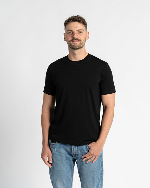 Un hombre con cabello corto y castaño oscuro y una barba bien recortada está de pie, centrado contra un fondo blanco liso, mirando directamente al espectador. Lleva una camiseta negra de cuello redondo y mangas cortas de la marca menique, que muestra el ajuste y la sencillez de la prenda. La camiseta está hecha de tejido de lana merina. También lleva unos vaqueros azul claro con corte recto y un bolsillo delantero visible, con una mano metida de forma casual en el bolsillo.