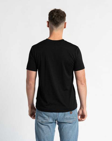 Vista trasera de un hombre que lleva una camiseta negra y vaqueros azul claro, de pie frente a un fondo blanco. La camiseta negra es el foco principal, mostrando su ajuste regular y diseño desde atrás. El hombre está situado en el centro, mostrando claramente la parte trasera de la prenda.