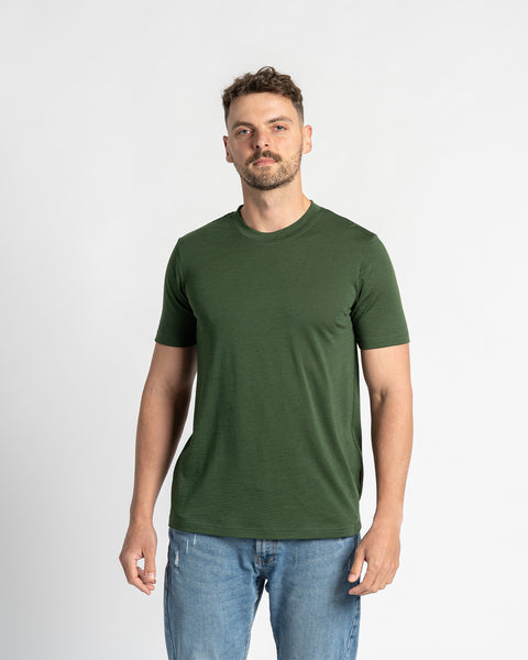 Un hombre modela una camiseta de cuello redondo de lana merina verde oscuro sobre un fondo blanco. La camiseta es el centro de atención de la imagen, mostrando su color y ajuste. También lleva puestos unos vaqueros de lavado claro.