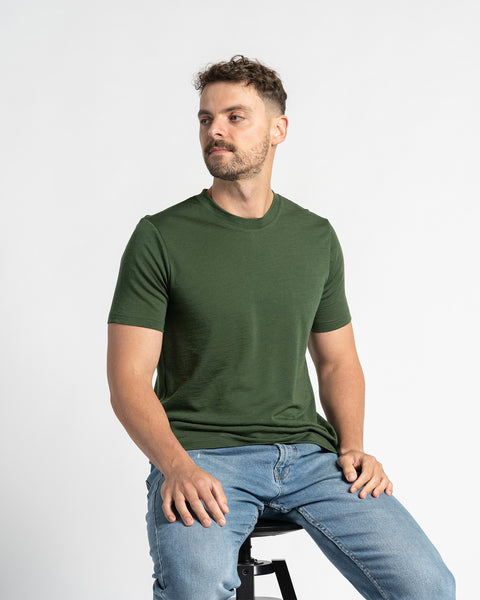 Hombre con una camiseta verde oscuro de Menique y jeans claros sentado en un taburete negro, mirando ligeramente hacia un lado, con un fondo blanco.