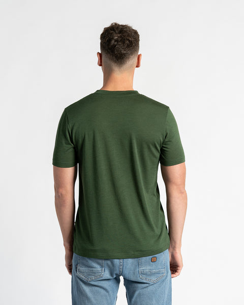 Vista trasera de un hombre con una camiseta verde oscuro de lana merina y unos vaqueros azul claro, de pie frente a un fondo blanco.