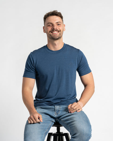 Un hombre que lleva una camiseta color mezclilla y unos vaqueros azul claro está sentado en un taburete contra un fondo claro. Tiene el cabello corto y castaño, una barba ligera y está sonriendo. La camiseta de lana merina de menique es el foco principal, mostrando su ajuste y apariencia mientras el hombre está sentado.