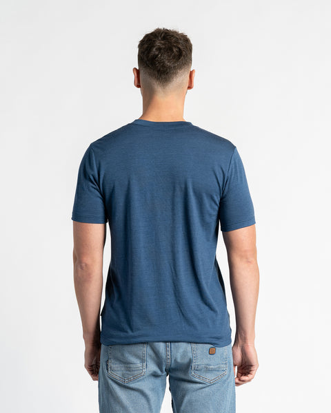 Vista trasera de un hombre que lleva una camiseta de lana merina Menique y vaqueros azul claro, de pie frente a un fondo blanco. La camiseta de color vaquero es el foco principal, mostrando su ajuste y diseño desde atrás. El hombre está colocado en el centro, mostrando claramente la parte trasera de la prenda.