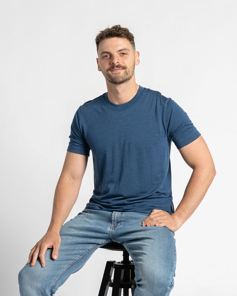 Un hombre que lleva una camiseta color mezclilla y unos vaqueros azul claro está sentado en un taburete contra un fondo claro. Tiene el cabello corto y castaño, una barba ligera, y sonríe con la mano apoyada en la cadera. La camiseta de lana merina es el foco principal, mostrando su ajuste y apariencia mientras el hombre está sentado.