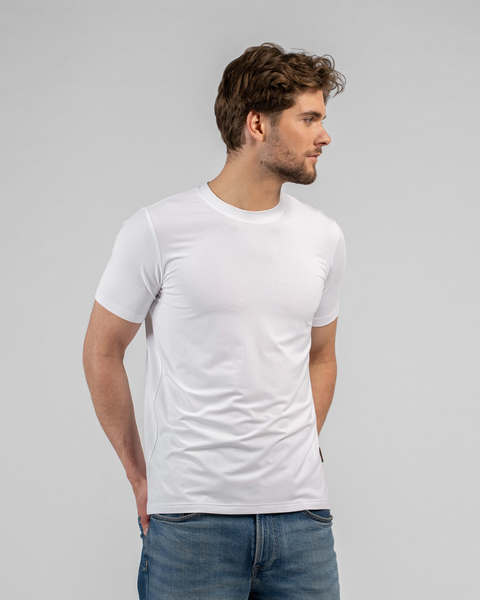 A young male model with brown hair and a light beard wearing a classic white short-sleeve t-shirt and blue jeans. He is standing against a light gray background, turned slightly to the side with his hands resting in his front pockets.