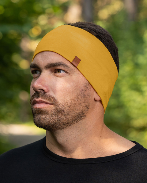 Un hombre con cabello corto y oscuro y barba aparece desde el pecho hacia arriba, llevando una cinta para la cabeza de lana merina color amarillo picante con una pequeña etiqueta marrón. Viste una camisa negra y mira hacia su izquierda con una expresión neutral. El fondo está suavemente desenfocado, mostrando follaje verde y troncos de árboles, lo que sugiere un entorno al aire libre.