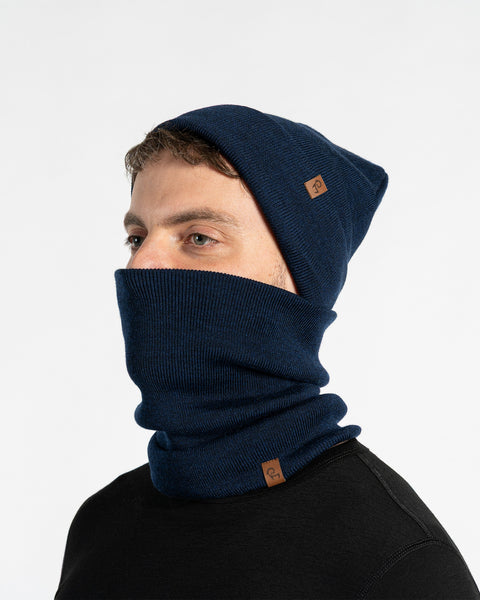 Un modelo masculino, visto de perfil, lleva un gorro azul y un cubrecuello a juego que le cubre la nariz y la boca. Tanto el gorro como el cubrecuello tienen un pequeño parche de cuero marrón con el logo de Menique. Tiene barba corta y cabello oscuro. El fondo es blanco sólido.