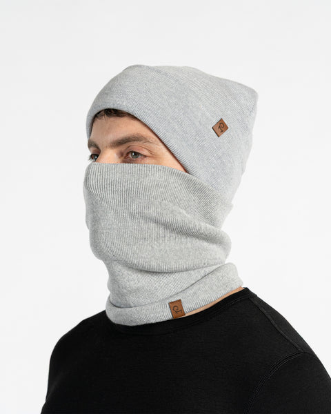 Un modelo masculino, visto de perfil, lleva un gorro de punto gris claro y un cubrecuello a juego que le cubre la nariz y la boca. Tanto el gorro como el cubrecuello tienen un pequeño parche de cuero marrón con el logo de Menique. Tiene barba corta y cabello oscuro. El fondo es blanco sólido.