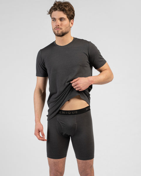 A studio portrait of a man standing and looking off to his left against a plain light grey background. He is wearing a charcoal grey short-sleeved t-shirt and matching long boxer briefs, gently pulling the t-shirt up with one hand to reveal the waistband.