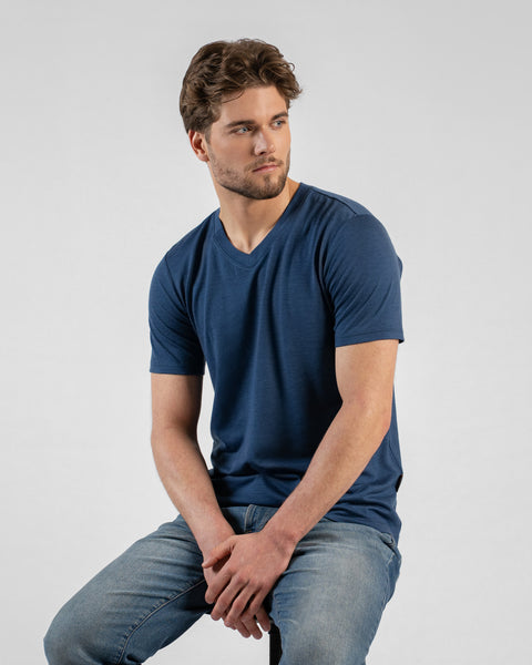 A studio portrait of a man sitting on a black stool, looking off to the right side of the frame with his hands clasped between his knees. He wears a denim blue v-neck t-shirt and blue jeans.