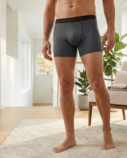 Un hombre de pie descalzo en una sala de estar luminosa y moderna, vistiendo calzoncillos bóxer gris oscuro con una pretina “MENIQUE”. Su torso está fuera de cuadro. La habitación tiene suelos de madera, una alfombra clara, una silla con una manta y una gran planta en maceta cerca de una ventana.