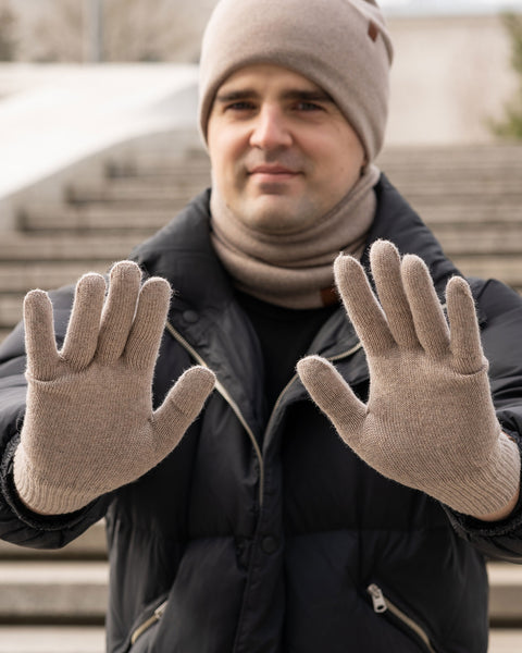 A man wearing a creamy beige knitted beanie and a matching neck gaiter, along with a dark puffer jacket, holds his hands up with palms facing the viewer, showcasing his menique gloves. He has a subtle smile and looks directly at the camera. The background is a blurred outdoor scene with stone steps.