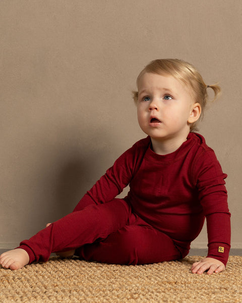 a girl sitting on the floor, wearing the Kids' Long Sleeve Set 160gsm Merino Wool thermal set in red color