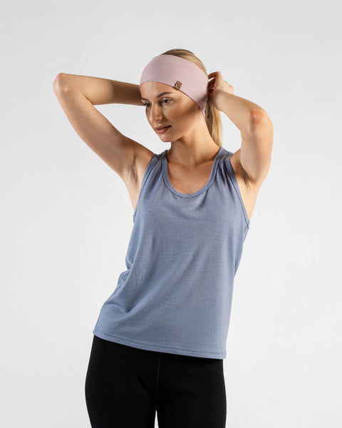 A woman standing against a white background, using both hands to adjust a dusty pink headband in her blonde hair. She is wearing a dusty blue sleeveless tank top and black leggings.