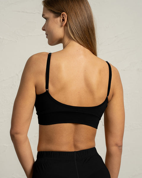 Una mujer vista de espaldas lleva un bralette negro de merino con tirantes finos y ajustables. La espalda baja y redondeada del bralette y la banda ceñida se ven claramente contra el fondo claro y texturizado.