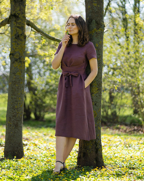 Una mujer de cabello castaño, vestida con un vestido morado de manga corta, está de pie al aire libre sobre el césped entre pequeñas flores amarillas.