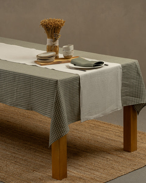 El mantel de lino menique en color verde a cuadros, cayendo sobre una mesa de madera, con un jarrón, una tabla de madera y un juego de platos para postre con pequeños cuencos de barro encima.