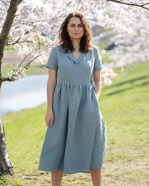 Una mujer de cabello castaño, vestida con un vestido cruzado de lino azul claro y mangas cortas, está de pie al aire libre junto a un árbol con flores blancas.