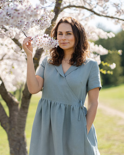 Una mujer de cabello castaño, vestida con un vestido cruzado de lino azul claro y mangas cortas, está de pie al aire libre junto a un árbol con flores blancas.