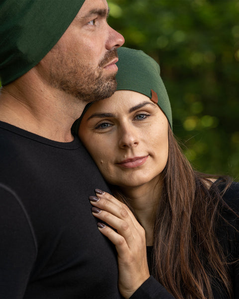 A picture featuring a couple wearing the unisex dark green merino beanie. It has a small logo on the side at the front. The woman is resting her head and palm on the man's chest. He is looking to the side.