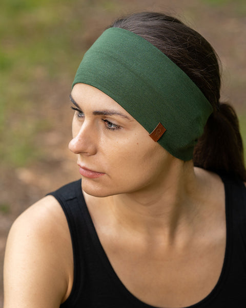 La imagen muestra a una mujer sentada en un bosque, vistiendo la cinta para la cabeza unisex menique de merino en verde oscuro. También lleva una camiseta sin mangas de merino negra. Ella mira hacia un lado.