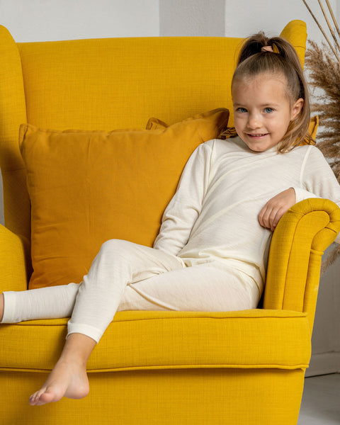 Una niña sentada en un sillón de color amarillo, vistiendo los pantalones para niños de Menique hechos de tela de merino en color natural.