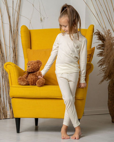 Una niña de pie junto a un sillón amarillo, sosteniendo un osito de peluche de la mano, vistiendo los Pantalones Merino 160 para Niños de menique en color Natural.