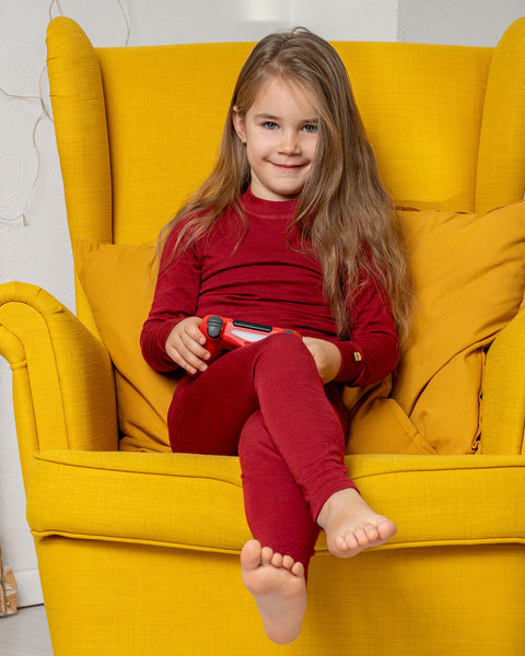 Una niña, sentada en un sillón amarillo, sosteniendo un mando de juego y vistiendo los Pantalones Merino 160 para niños de menique en color Cereza Real.