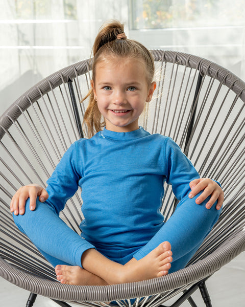 Una niña sentada en una silla de descanso que lleva puestos los pantalones Menique Kids 160 Merino en color azul claro. Tiene las piernas cruzadas y las palmas de las manos sobre las rodillas.