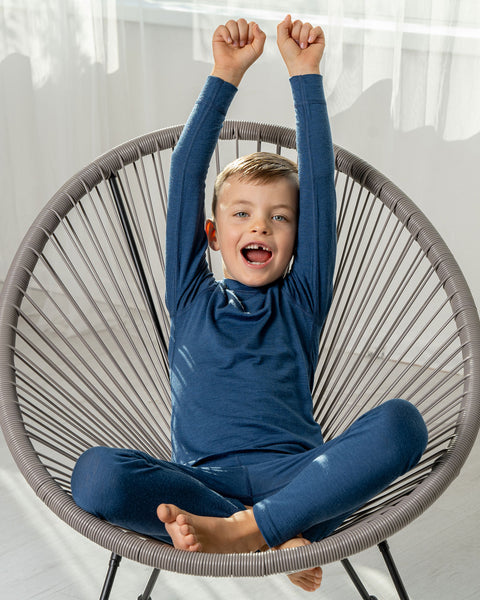 A boy sitting in a lounge chair, wearing the menique Kids' Merino 160 Long Sleeve Set in Denim color.