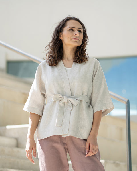Una mujer de cabello castaño, vestida con una chaqueta estilo kimono color beige claro sobre pantalones malva, está de pie en unas escaleras interiores de concreto.