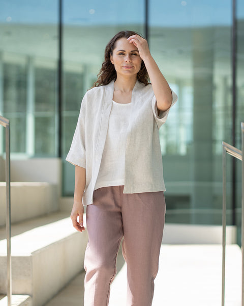 Una mujer de cabello castaño, vestida con una chaqueta menique natural de estilo kimono sobre pantalones malva, está de pie en unas escaleras interiores de concreto.