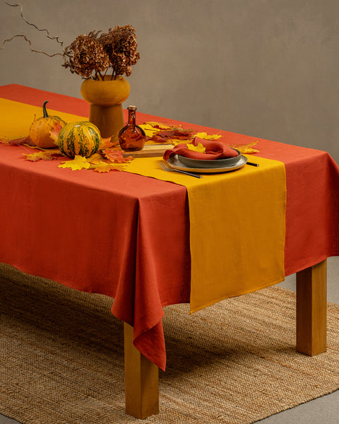 El mantel de lino menique en color rojo canela, cayendo sobre una mesa de madera, con un jarrón encima.