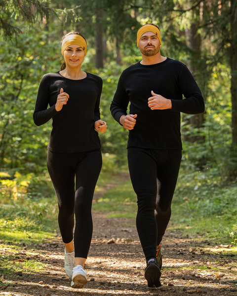 Un hombre y una mujer están trotando hacia el espectador por un sendero de tierra a través de un bosque salpicado de sol. Ambos llevan cintas para la cabeza de un amarillo intenso, camisetas negras de manga larga y mallas negras, lo que sugiere que podrían ser una pareja haciendo ejercicio juntos.