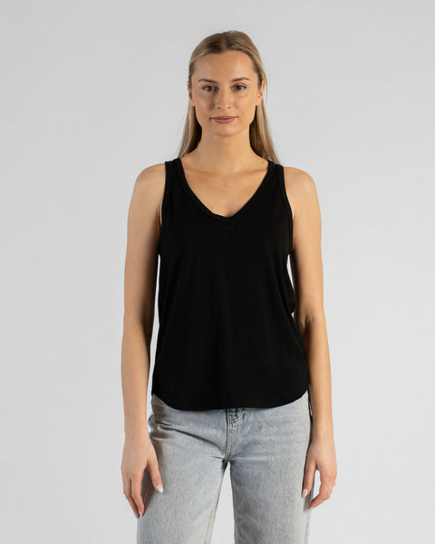 A studio portrait of a blonde woman standing and looking directly at the camera with her arms relaxed by her sides. She is wearing a regular fit, solid black sleeveless v-neck top and light-wash blue jeans against a plain light grey background.
