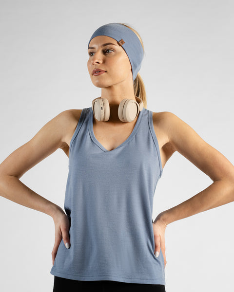 A woman stands against a light grey background with her hands on her hips, looking off to the side. She wears a dusty blue sport tank top, a matching dusty blue headband, and beige headphones resting around her neck.