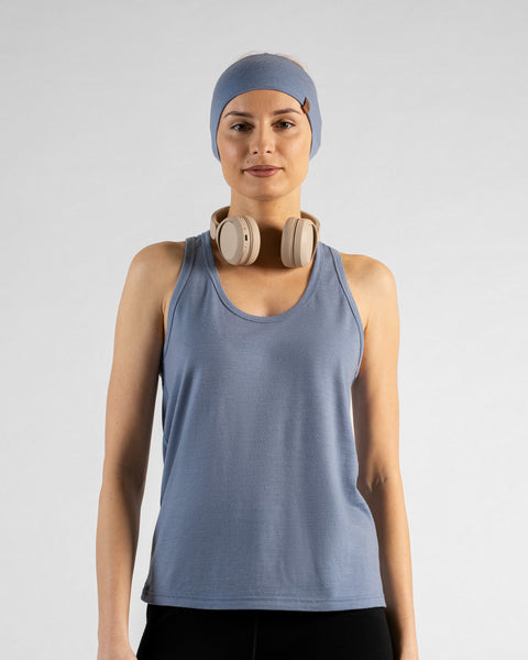 A profile shot of a woman standing against a white background, wearing a dusty blue sleeveless top with a small brown logo tag on the side and black leggings. Her blonde hair is pulled back in a ponytail, and she is gently touching her face with one hand.