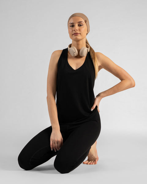 A woman kneeling on a light grey background, wearing a black sleeveless top, black leggings, and a beige headband. She has beige headphones resting around her neck, her left hand is on her hip, and she is barefoot.