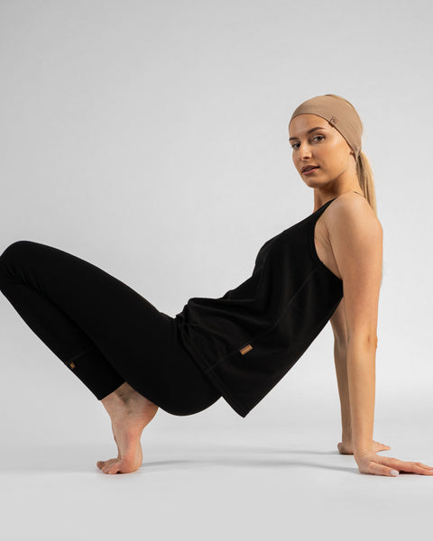A barefoot woman sitting on a light grey background, leaning back and supporting her weight on her hands with her arms extended behind her. She has her knees bent and legs lifted, balancing playfully on the toes of her left foot. She is wearing a black sleeveless top, black leggings, and a beige headband.