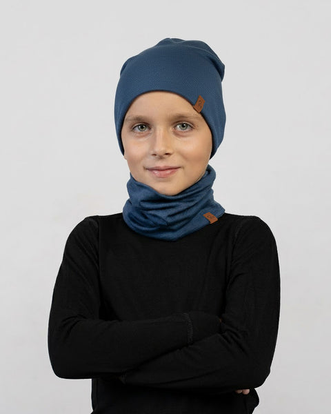 Un niño lleva un conjunto de gorro y braga de cuello azul oscuro con una camiseta de manga larga negra, posa con los brazos cruzados y una leve sonrisa frente a un fondo gris claro.