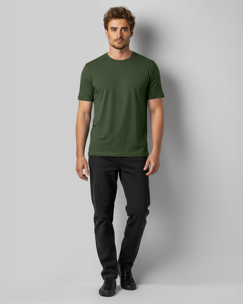 Un hombre está de pie mirando hacia adelante, vistiendo una camiseta verde, vaqueros negros y zapatillas negras sobre un fondo gris liso.