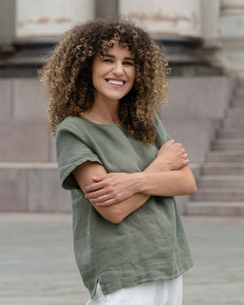 Hermosa mujer con cabello rizado en la ciudad, posando con una blusa de lino verde piedra Emma y pantalones de lino blanco puro Dakota.
